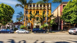 A multi-story building with a yellow facade, palm trees, and parked cars on the street.