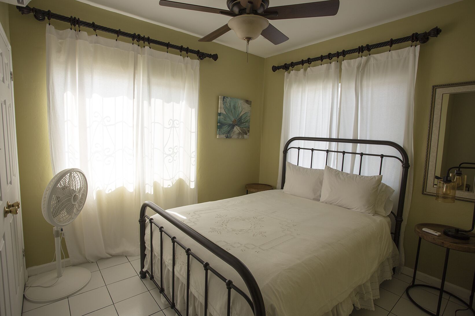 Quarto com cama, ventoinha no teto, mesa de cabeceira e janela com cortinas.