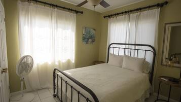 Quarto com cama, ventoinha no teto, mesa de cabeceira e janela com cortinas.