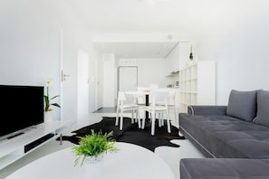 A modern living room with a flat-screen TV, a dining area with a white table and chairs, and a grey sofa.