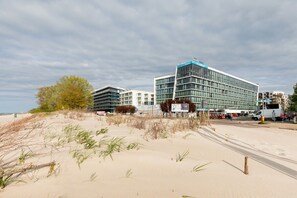 Een hotel aan de kust met een moderne glazen gevel, nabij een zandstrand met weinig begroeiing.
