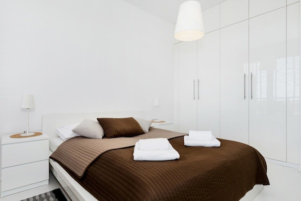 A modern bedroom with a large bed, white nightstands, a hanging light fixture, and a wardrobe.