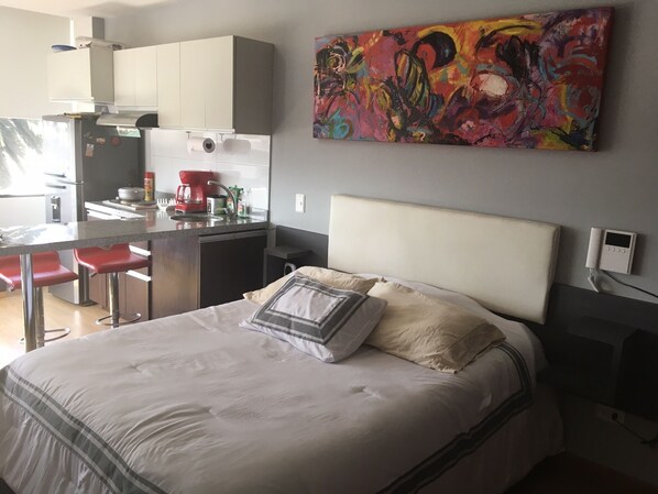A modern bedroom with a large bed, a small kitchenette, and a colorful abstract painting on the wall.