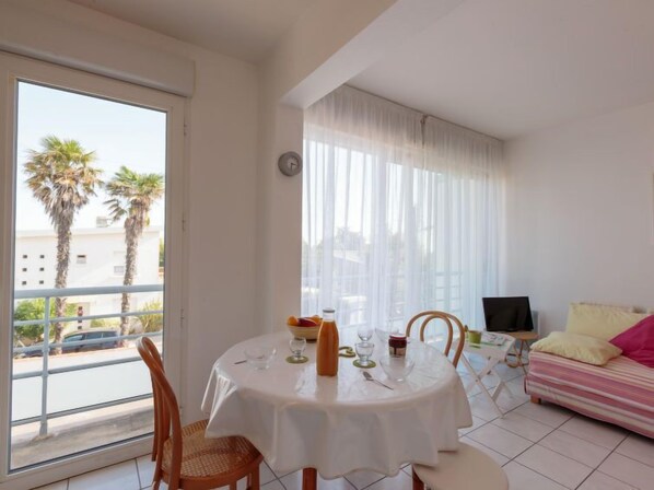 A room with a dining table set for two, a sofa, a TV, and a balcony with palm trees.