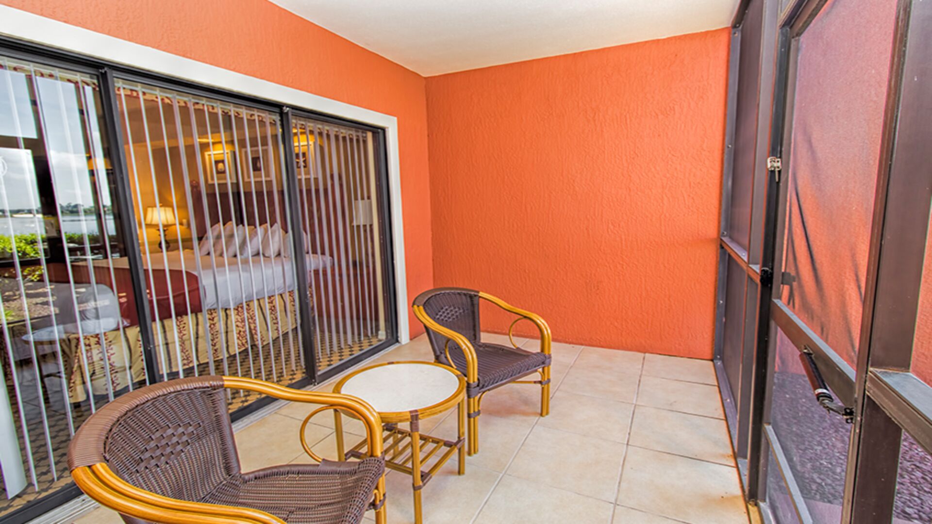 Ein Balkon mit zwei Korbstühlen und einem kleinen Tisch, eine Glasschiebetür und ein Bett mit weißem Kopfteil, das durch die Tür sichtbar ist.