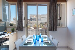 A dining area with a table set for two, a kitchen area with a stove, and a view of the neighborhood through large windows. - Aretusa Apartment with Terrace by Wonderful Italy (Siracusa)