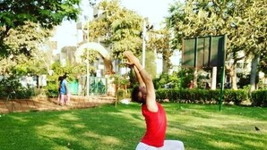 Una persona realizando una postura de yoga sobre una colchoneta morada en un parque con césped y árboles.