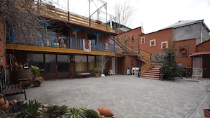 Un patio con suelo de piedra, plantas en macetas, una fuente pequeña y un edificio con balcón y escaleras.