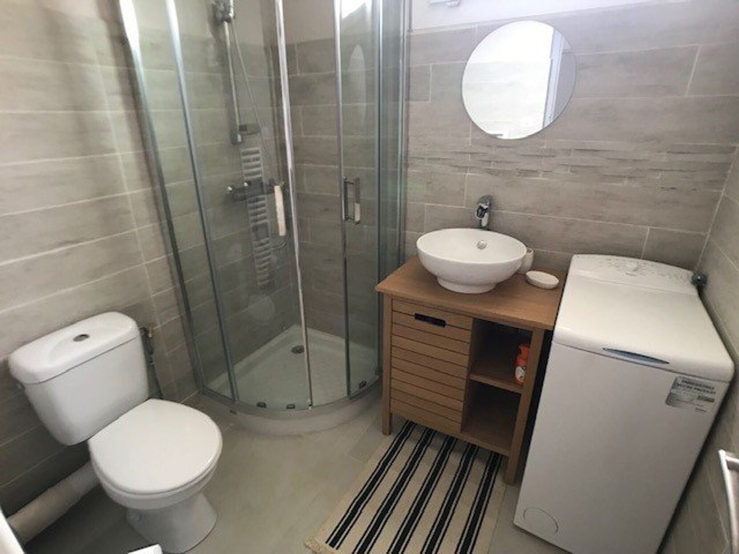 Ein modernes Badezimmer mit Duschkabine aus Glas, einer weißen Toilette, einem Waschbecken mit Spiegel darüber und einem kleinen, weißen Toilettenpapierhalter.
