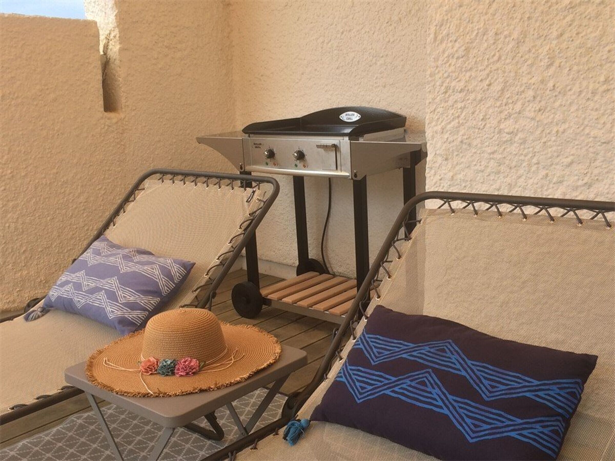 A sun lounger with a woven hat and patterned pillows, a small grill, and a metal grill stand against a textured wall.