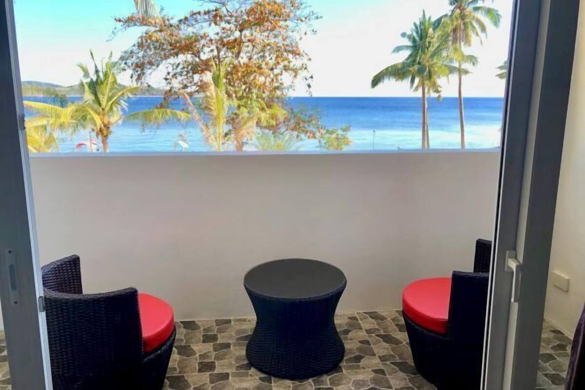 Een balkon met twee rieten stoelen en een ronde tafel, met uitzicht op een strand met palmbomen en de oceaan.