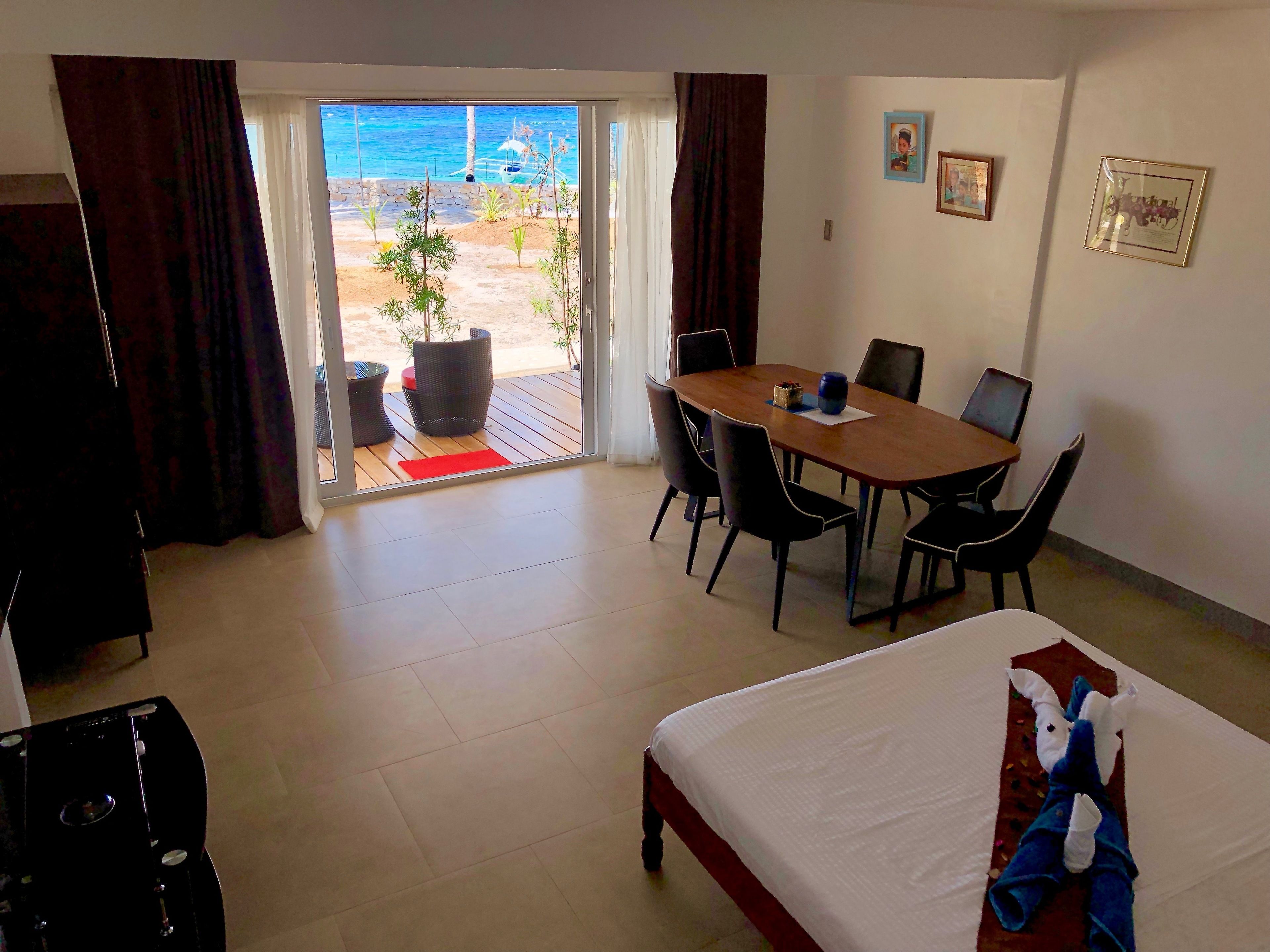 A hotel room with a bed, a dining table with chairs, a view of the beach, and a balcony with potted plants.