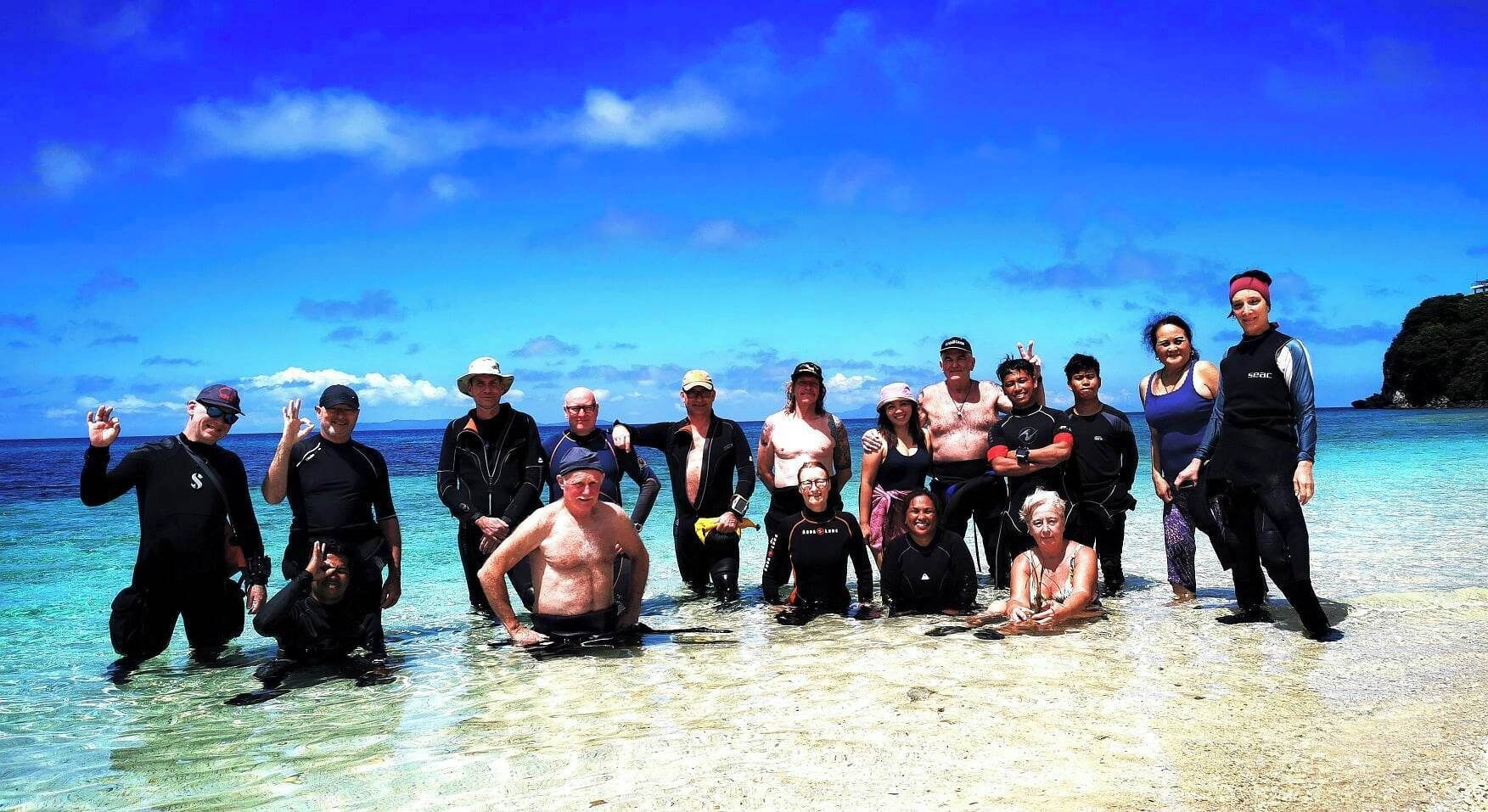 Eine Gruppe von Menschen steht an einem Strand mit klarem, blauem Wasser und einer felsigen Landzunge im Hintergrund.