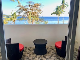Un balcón con sillas de mimbre y una mesa, con vistas a una playa con palmeras y el océano.