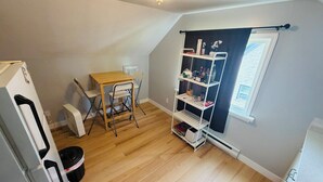 Een kleine, goed verlichte kamer met houten vloer, een tafel met stoelen, een witte plankkast en een raam met uitzicht.