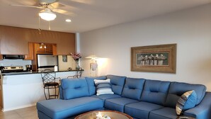 Ein Wohnzimmer mit einem blauen Ecksofa, einem runden Couchtisch und einer Küchenzeile mit Holzausstattung und einer Mikrowelle.
