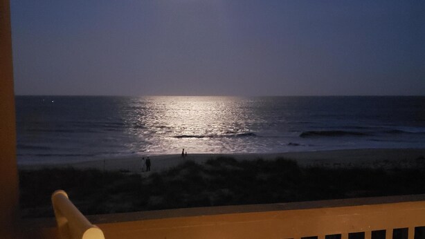 Eine Holzterrasse mit einem Stuhl, mit Blick auf einen Strand in der Nacht, bei sichtbarem Mond und Meereswellen.