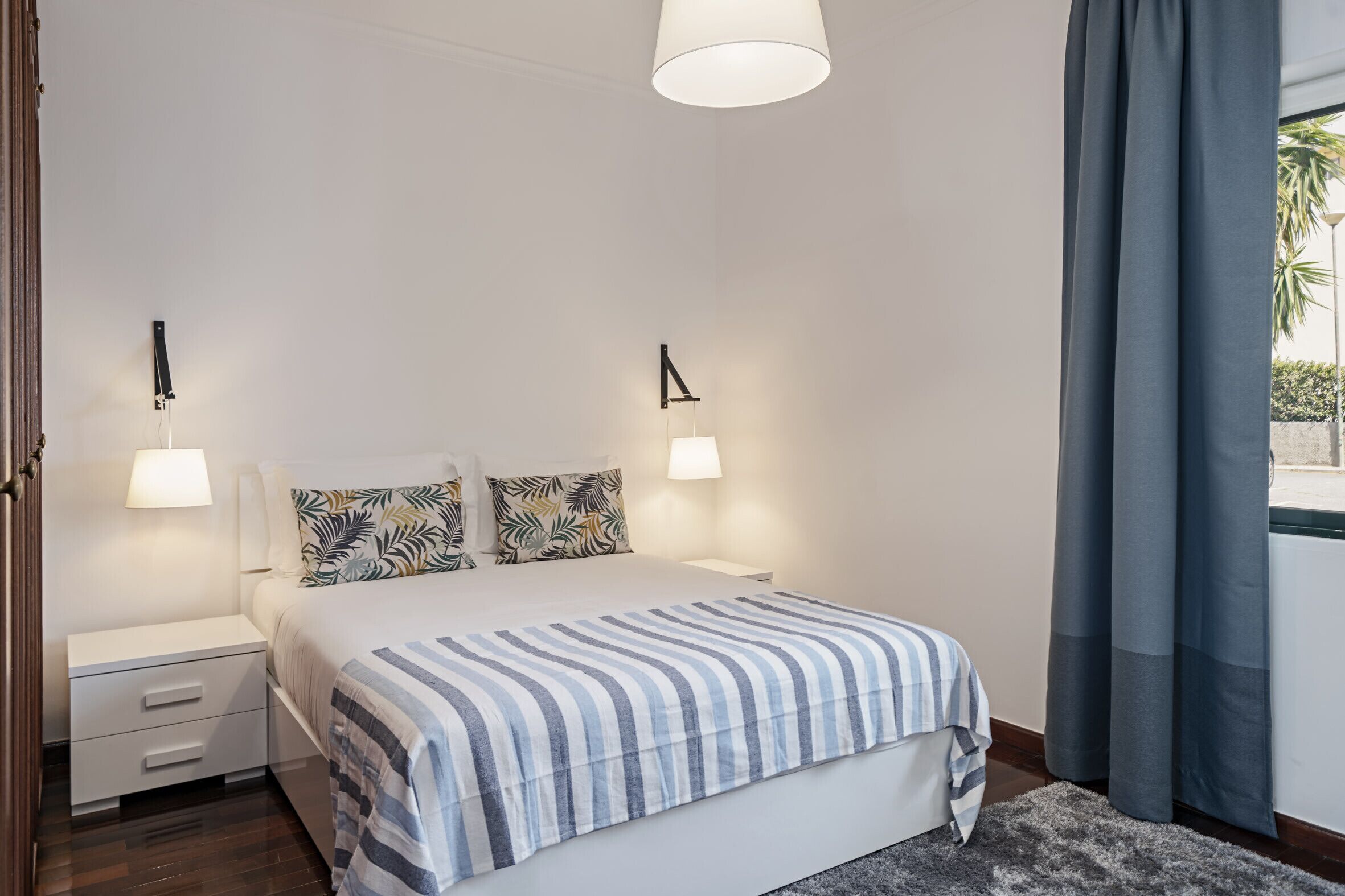 A bedroom with a bed, two bedside tables, a nightstand, a lamp, and a window with curtains.