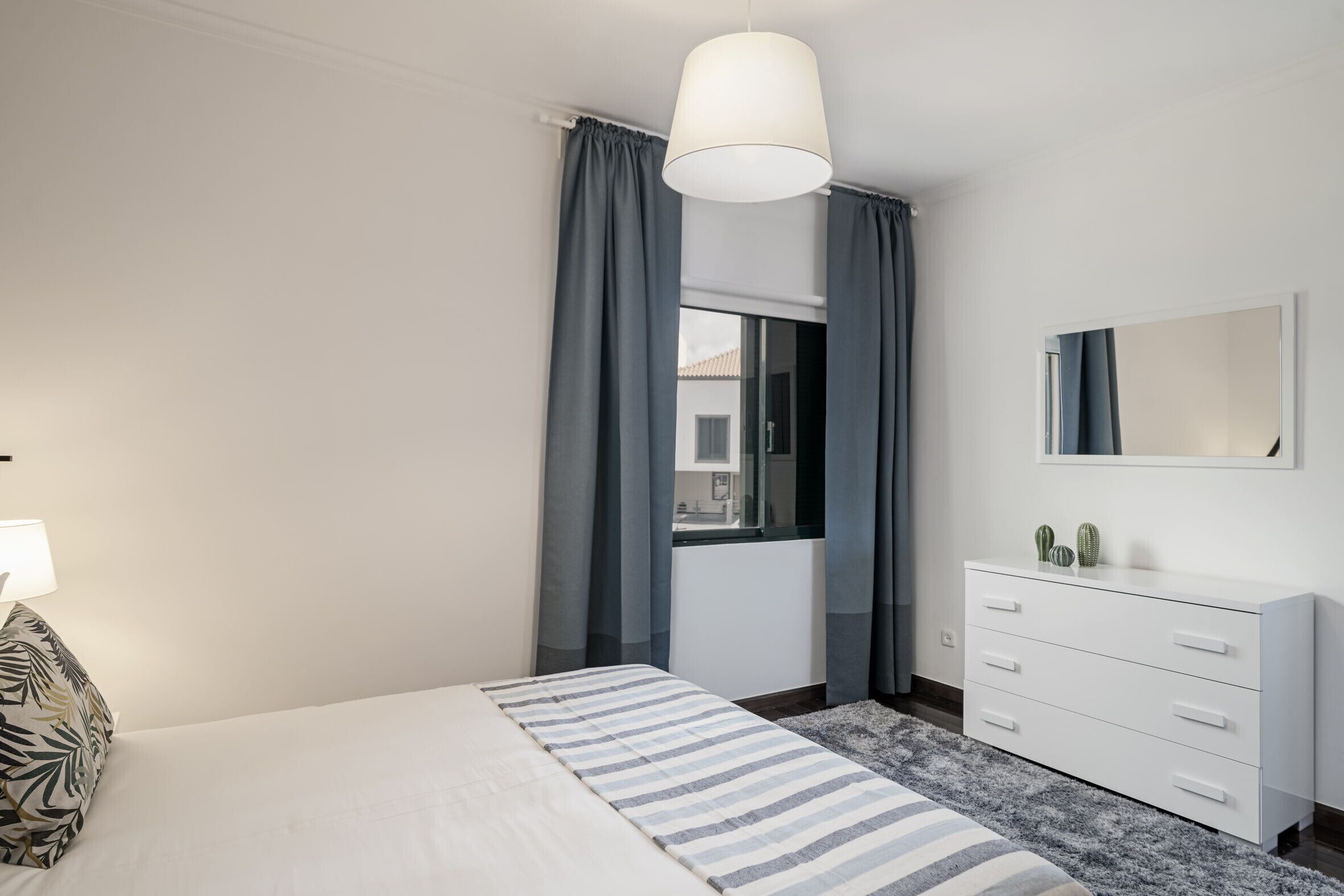 A modern bedroom with a bed, a dresser, a mirror, and a window with curtains.