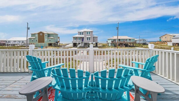 Una terraza con sillas Adirondack azules y una mesa, con vista a una playa con casas y una cerca blanca de piquetes.