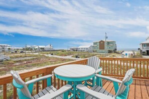 Une terrasse avec une table ronde et des chaises, donnant sur une plage et des bâtiments.