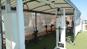Ein überdachter Außenbereich für Fitnessgeräte, darunter ein Squat-Rack und Gewichtsbänke.