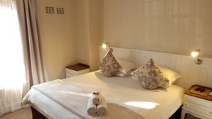 A neatly made bed with a rolled towel, two decorative pillows, a bedside table with a lamp, and a nightstand with a book and remote controls. - 137 High Street Guest House (Makhanda)