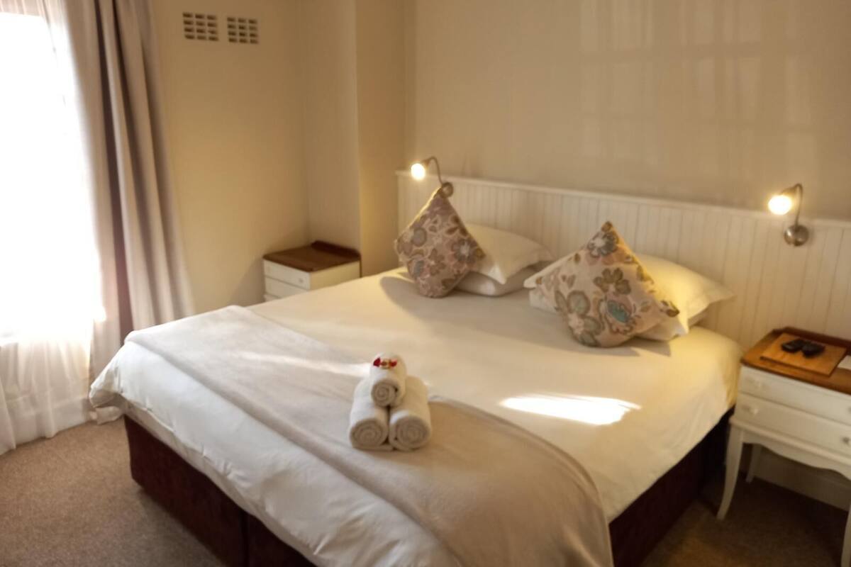 A neatly made bed with a rolled towel, two decorative pillows, a bedside table with a lamp, and a nightstand with a book and remote controls.