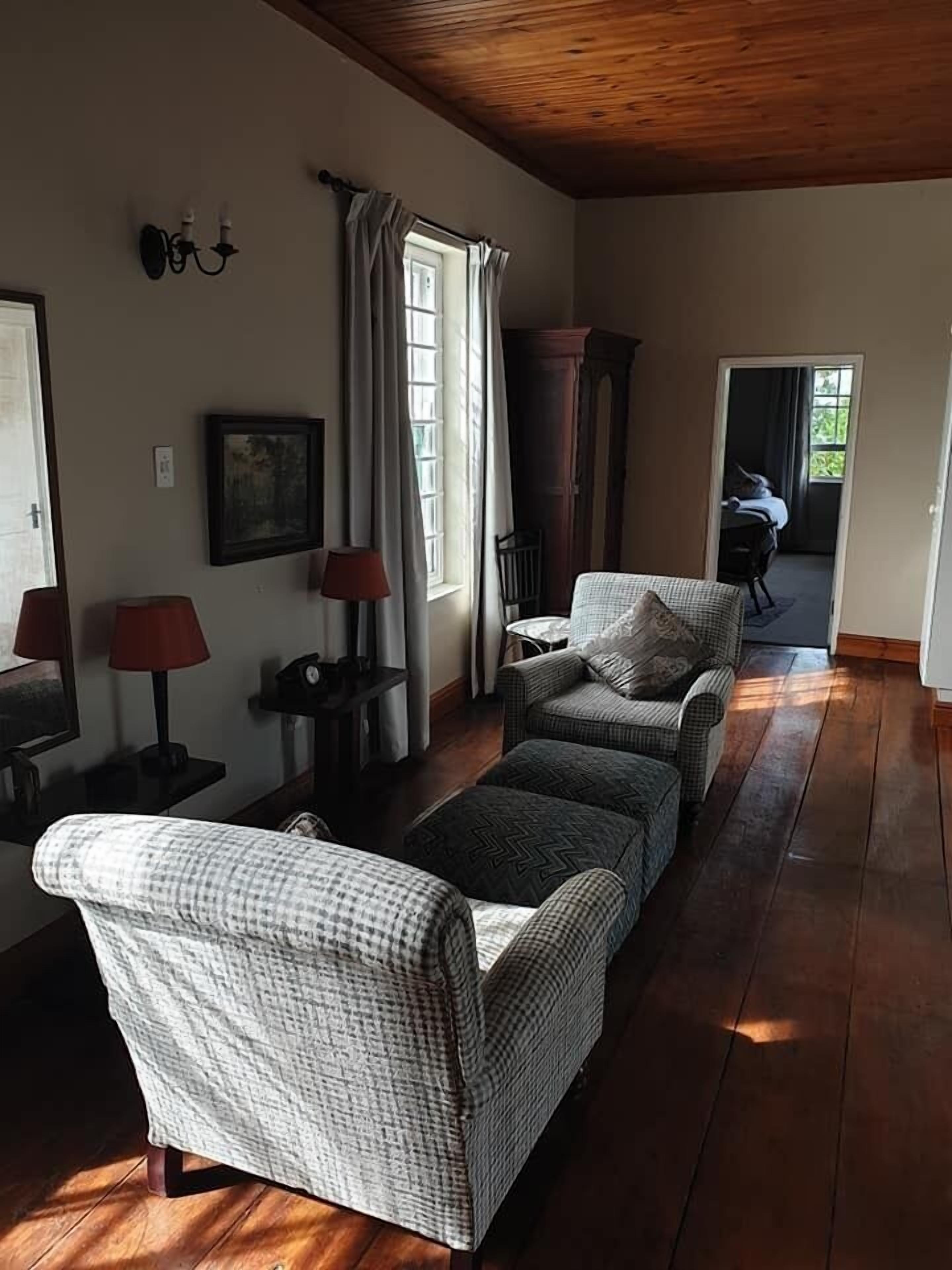 Una sala de estar acogedora con un sofá, un sillón y piso de madera. Se ve una ventana con cortinas, una lámpara y un cuadro en la pared.