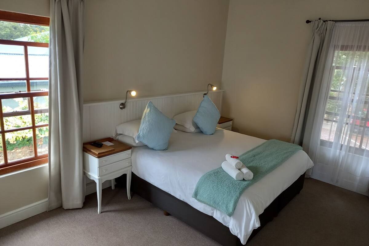 A neatly made bed with a white headboard, two pillows, and a teal throw blanket. There is a bedside table with a lamp and a remote control. The room has beige walls and a large window with curtains.