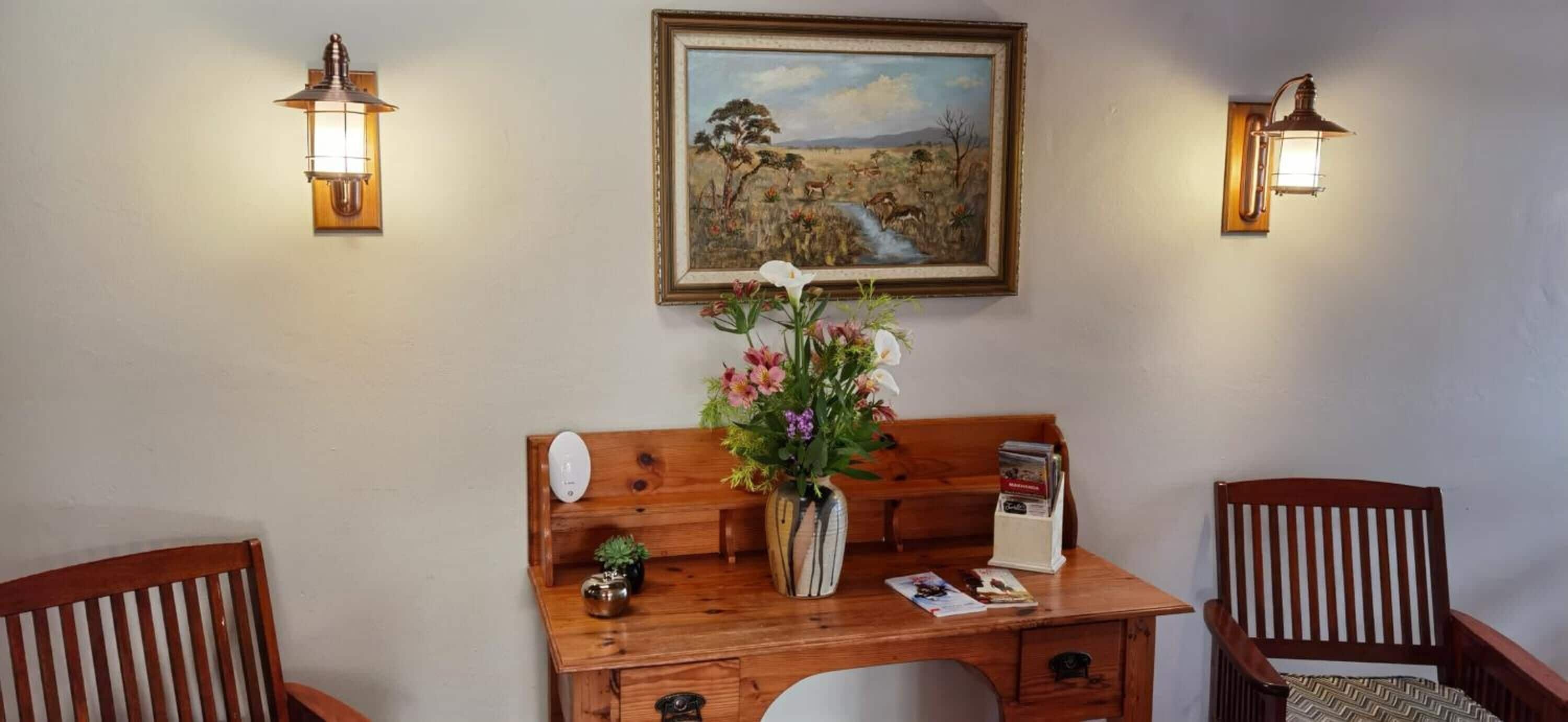 A wooden desk with a vase of flowers, a lamp, and a framed painting on the wall.