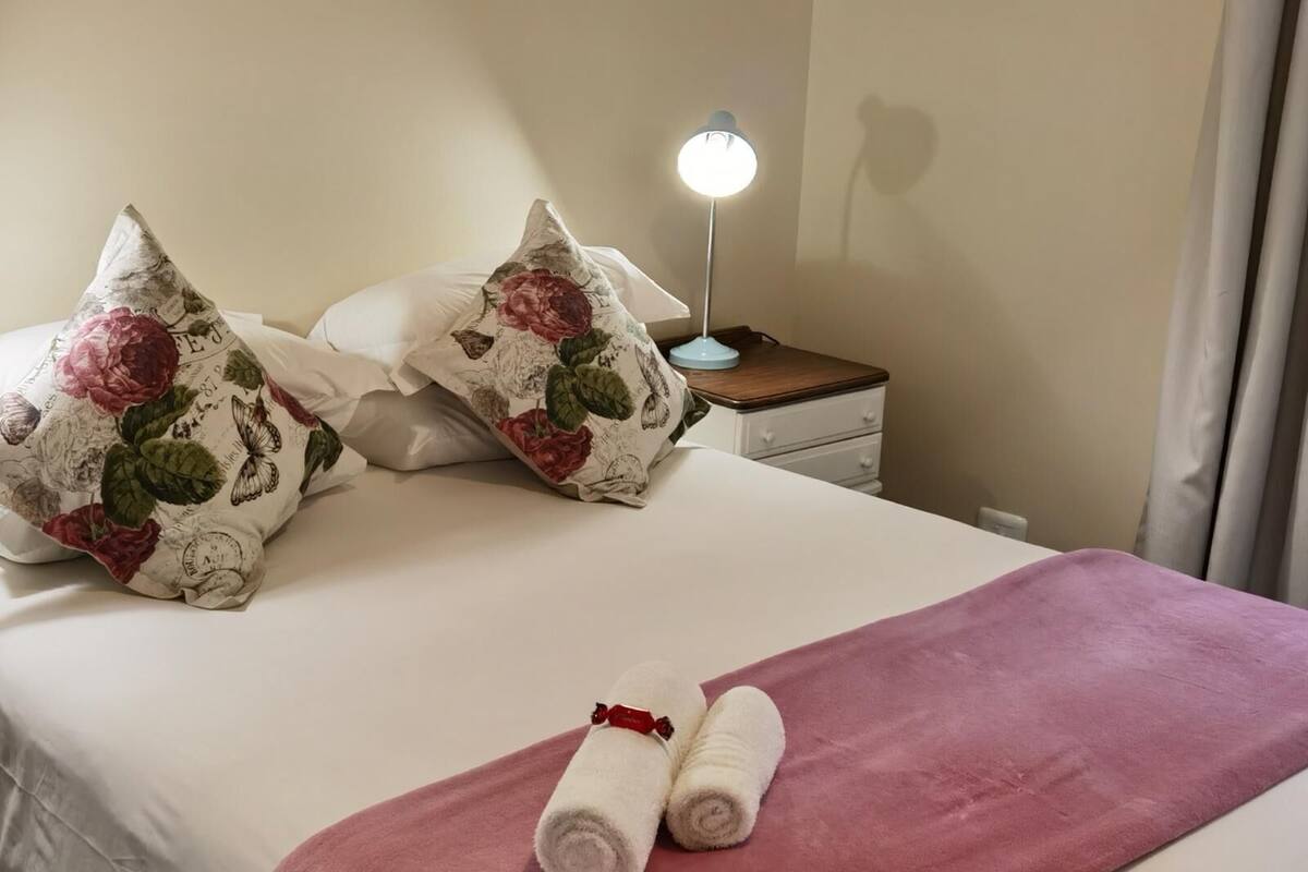 A neatly made bed with floral pillows, a pink blanket, and rolled towels.
