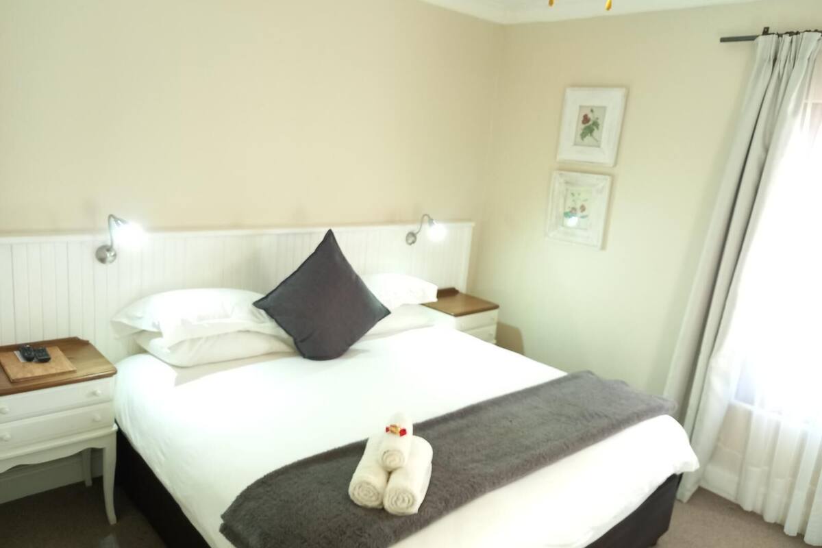 A neatly made bed with a dark pillow, white bedding, and a rolled towel on the footboard. There are two bedside tables with lamps, a framed picture on the wall, and a window with curtains.