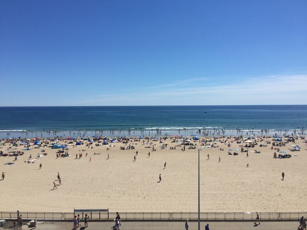 Ein Strand mit breiter Promenade, Sonnenschirmen und Menschen, die das Meer genießen.