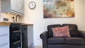 Ett modernt kök med en inbyggd ugn och diskmaskin, en svart lädersoffa med en enda kudde, en väggklocka och en stor abstrakt målning.