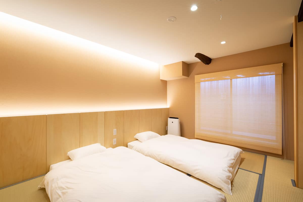 Une chambre d’hôtel avec deux lits, une grande fenêtre avec des stores et un luminaire au plafond.