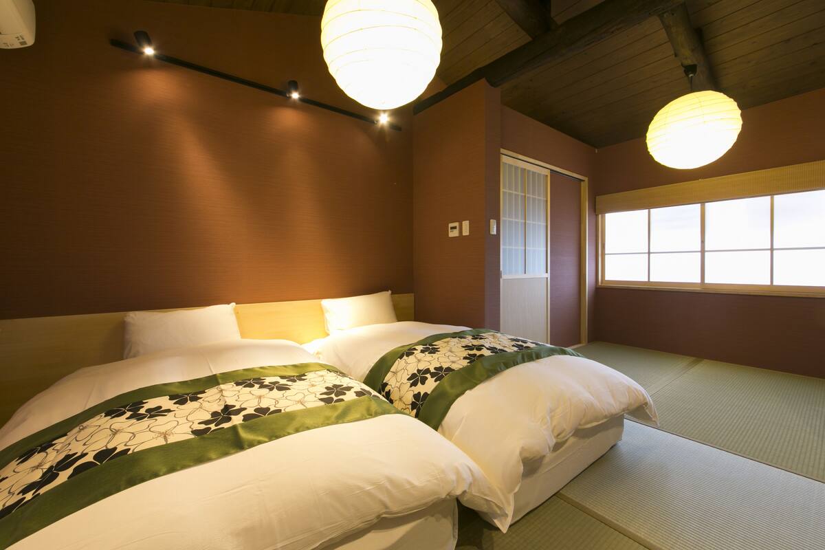 Ein Hotelzimmer mit zwei Betten, traditionellem japanischen Tatami-Bodenbelag, Holzbalkendecke und einem großen Fenster mit Aussicht.