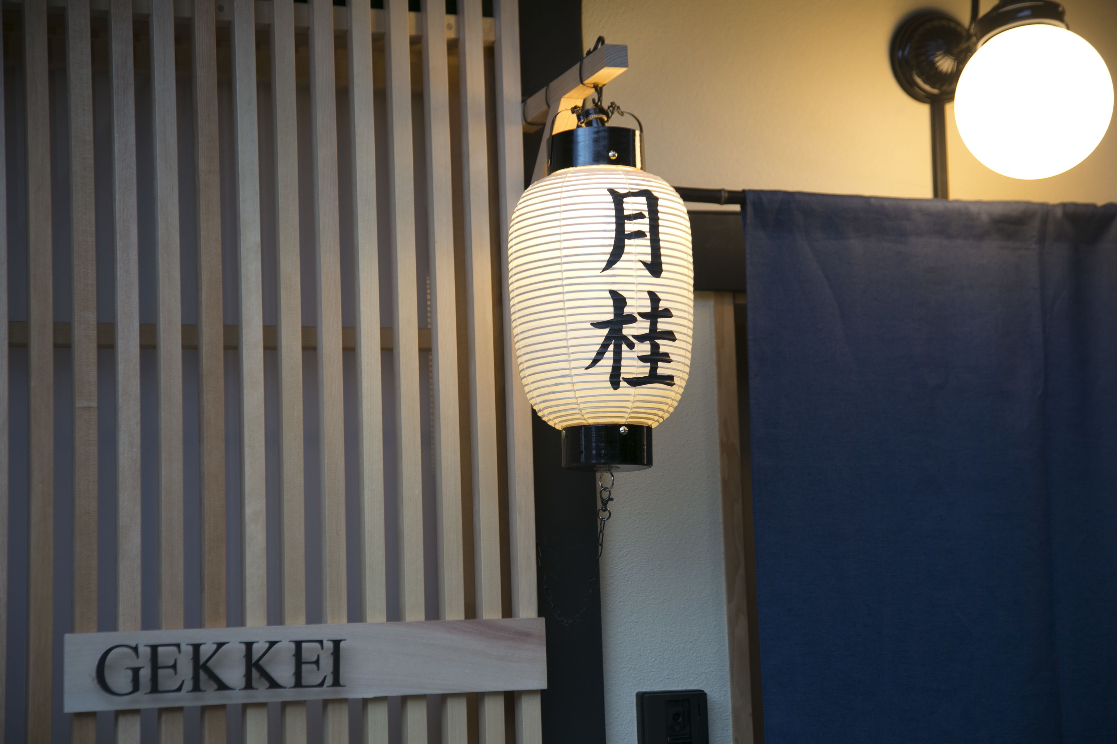 Un farol tradicional japonés con caracteres japoneses, un letrero que dice "GEKKEI" y una cortina azul al fondo.