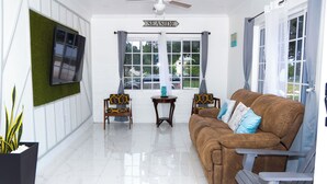 A living room with a brown sofa, two armchairs, a ceiling fan, a TV mounted on a green wall, and a window with curtains. - Treasure Beach Inn and Bar Room #1: Driftwood Queen (St. Elizabeth Parish)