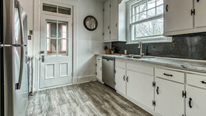 A kitchen with white cabinets, a stainless steel refrigerator, a wooden floor, a window with a view, and a clock on the wall.