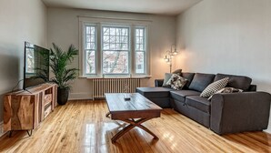 A modern living room with a sectional sofa, a wooden coffee table, a TV on a wooden stand, and a large window with a view of buildings.