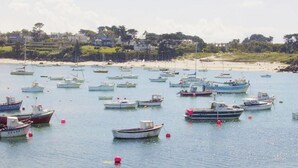 Eine Küstenansicht mit zahlreichen Booten, die in ruhigem Wasser vor Anker liegen, einem Sandstrand und Häusern im Hintergrund.