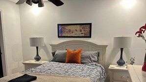 A bedroom with a bed, two bedside tables, a ceiling fan, a framed picture on the wall, and a nightstand with a lamp. - Downtown-Melbourne Beach-Dining-Cafes Walk to Everything (Melbourne)