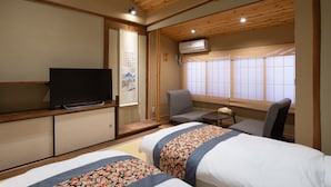 Quarto de hotel com vigas de madeira no teto, televisão sobre um móvel, duas camas com roupa de cama floral, uma cadeira, uma mesinha e uma janela com vista.
