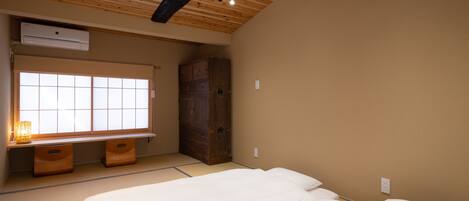 A bedroom with a bed, a window with blinds, a wooden wardrobe, and a small table with a lamp.