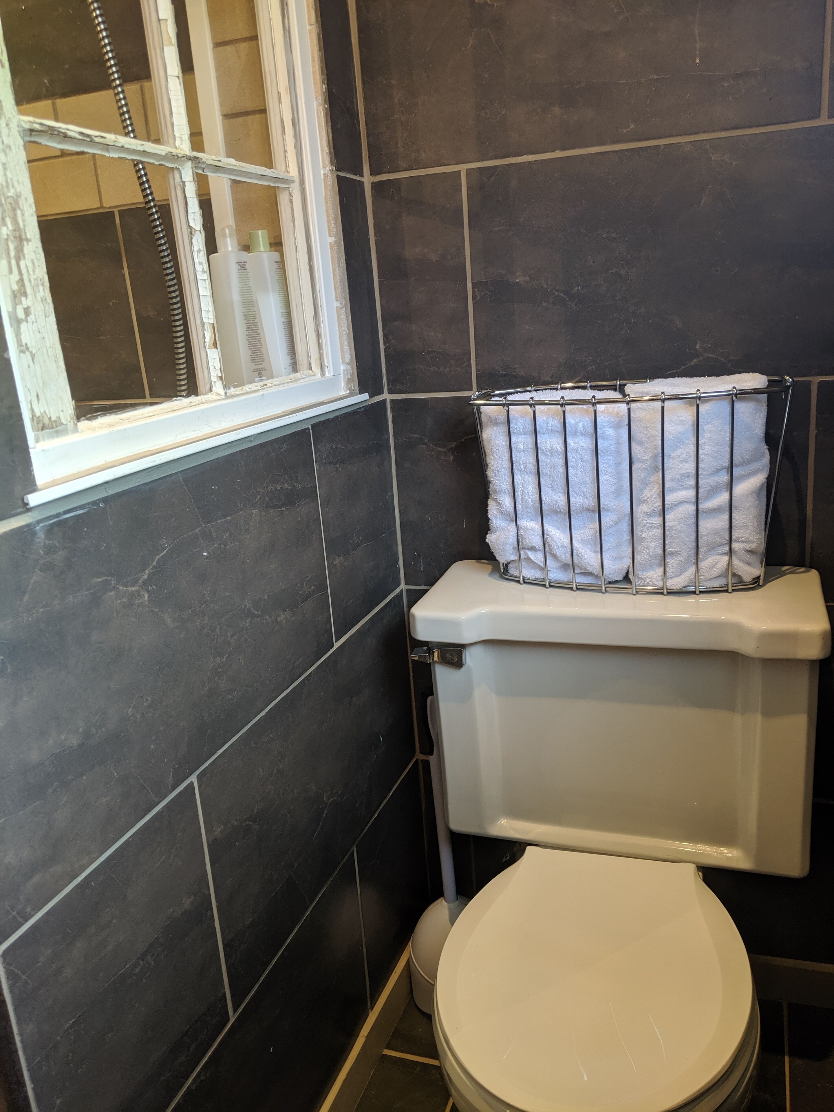 Un baño con azulejos oscuros, un inodoro blanco y una ventana con reja de metal.