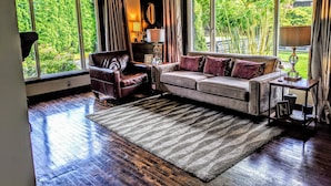Un salón con un sillón de cuero, un sofá con cojines estampados, una alfombra y ventanas amplias con vistas al verdor.