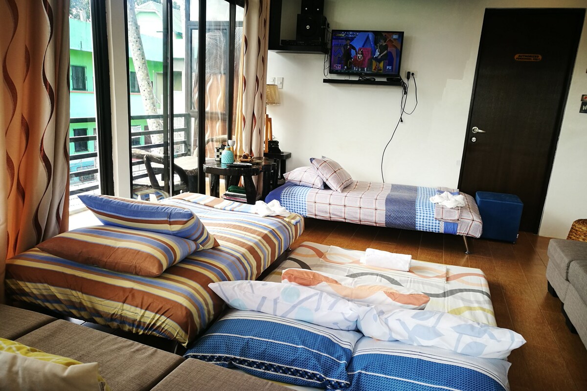 A room with a sofa, two beds with striped bedding, a television on the wall, and a view of buildings outside.