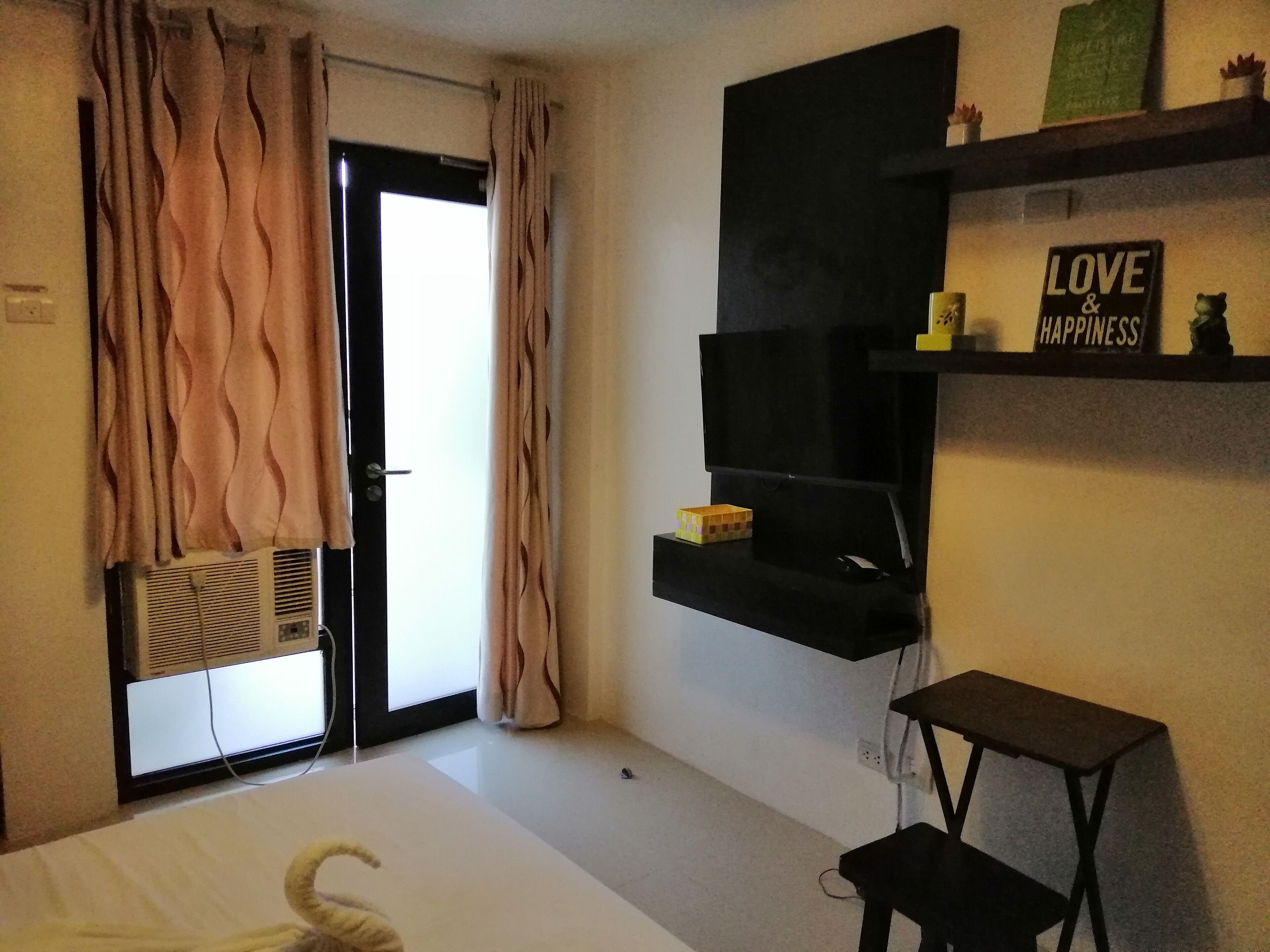 Una habitación con una cama, un televisor montado en la pared, una mesita y un estante con un cartel que dice "LOVE & HAPPINESS".