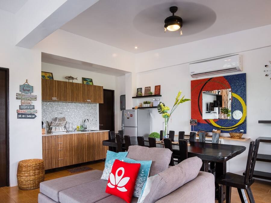 A modern living room with a sofa, a dining table, and a kitchen area with wooden cabinets and a tiled backsplash.